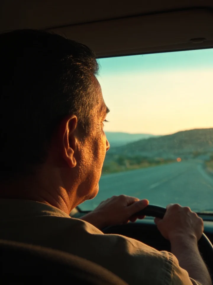 A man drives home at sunset; the low sun comes in through the windshield and the road opens out toward some hills.